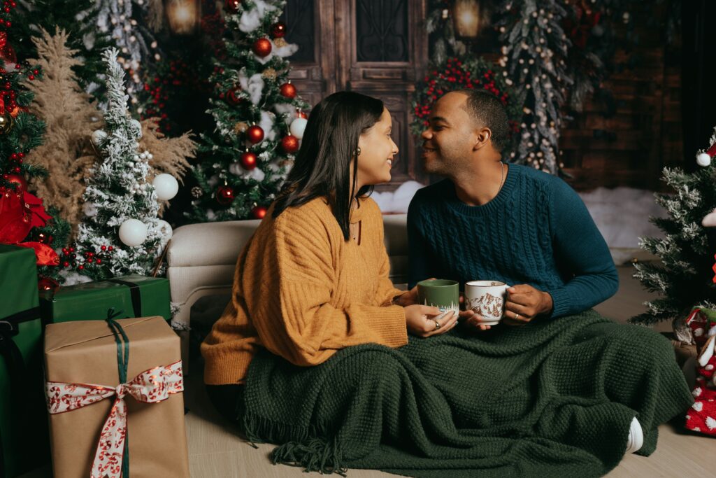 Female and male cuddled under blanket holding mugs with Christmas tree decorations in the background
